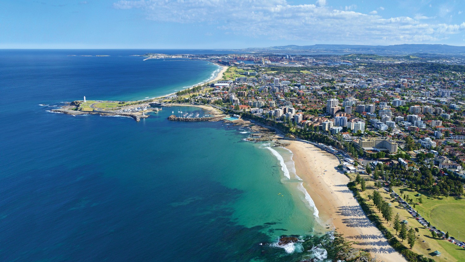 Aerial view of Wollongong’s coastline, showing beaches, the lighthouse, marina and city skyline beside the ocean.