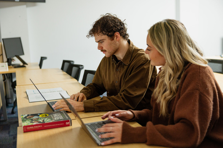 two students at desk with laptops open in front