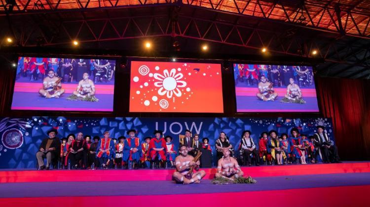 People sitting on stage during Acknowledgment to Country with three screens overhead