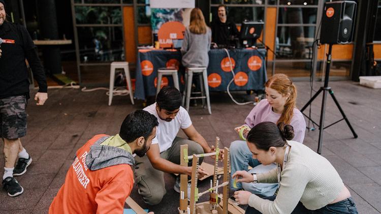 Four people sitting outside on a paved area building a structure with various items. One people is holding a wheel of tape.