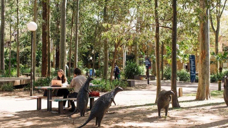 Two people are sitting on an outside table and bench with green trees surrounding them and three brass statues of Kangaroos in the foreground,