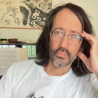 Professor Abe Roth with shoulder-length dark hair and rectangular glasses, wearing a light-colored T-shirt. The person is seated indoors with one hand resting against the side of the head. In the background, there is a piano or keyboard with sheet music, several small bowls or decorative objects on a shelf, a window, and a framed abstract artwork on the wall.