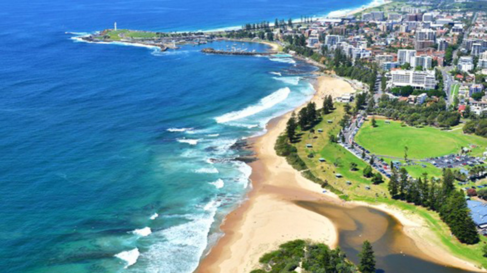 Aerial view of a coastal landscape featuring sandy beaches, vibrant blue waters, and a city skyline with green parks and a lighthouse.