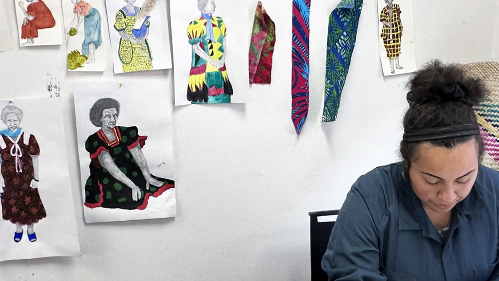 A person sits at a table surrounded by colorful fabric swatches and illustrated fashion designs displayed on the wall.
