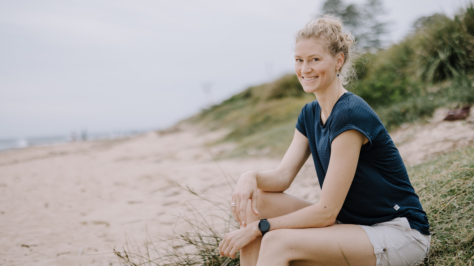 Dr Katharina Peters sits in the sand dunes on the beach. The water is in the background. She wears a blue top and beige shorts. Photo: Michael Gray