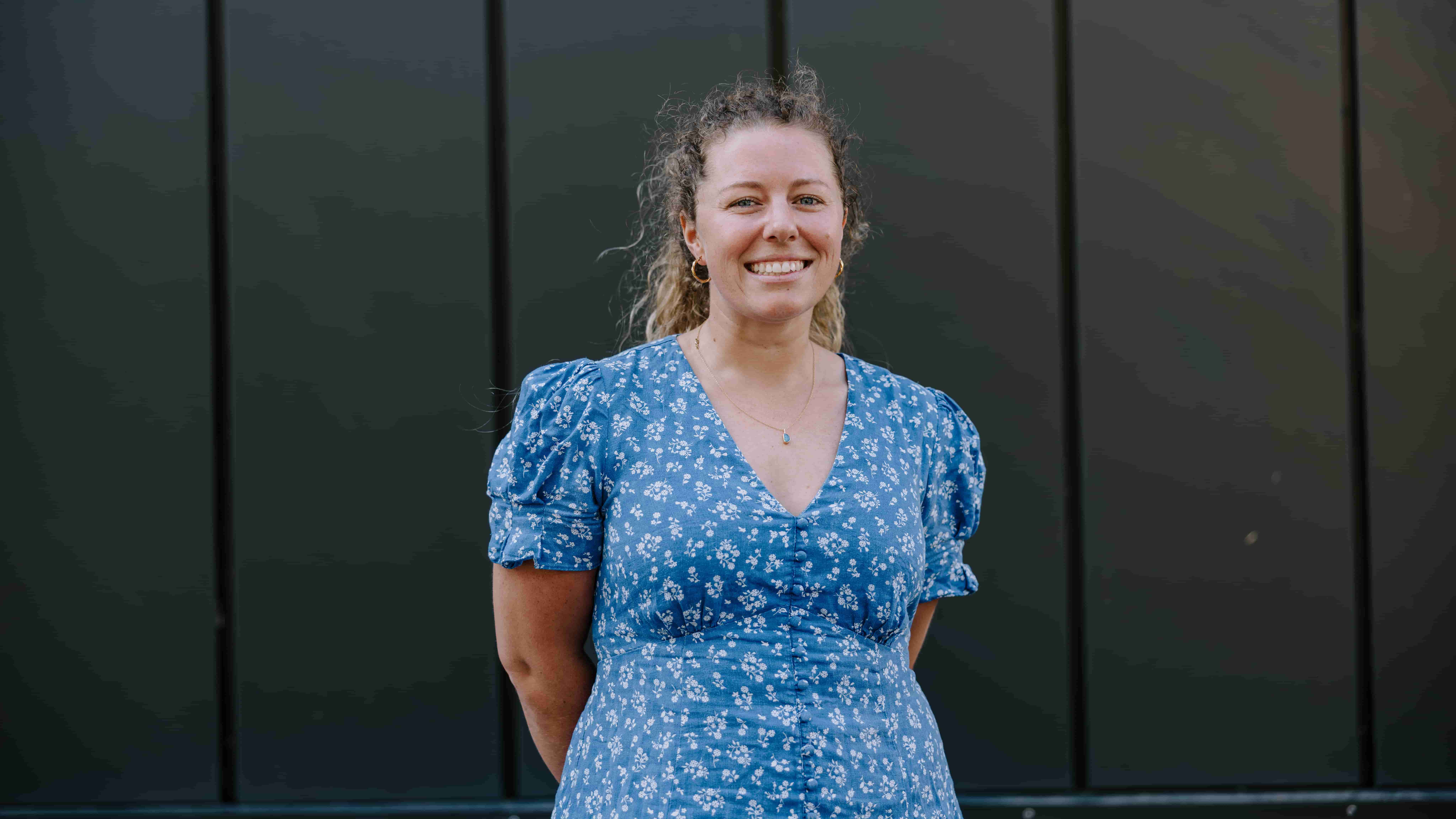 Dr Samara Walpole wears a blue dress and is smiling. She stands against the dark background of UOW