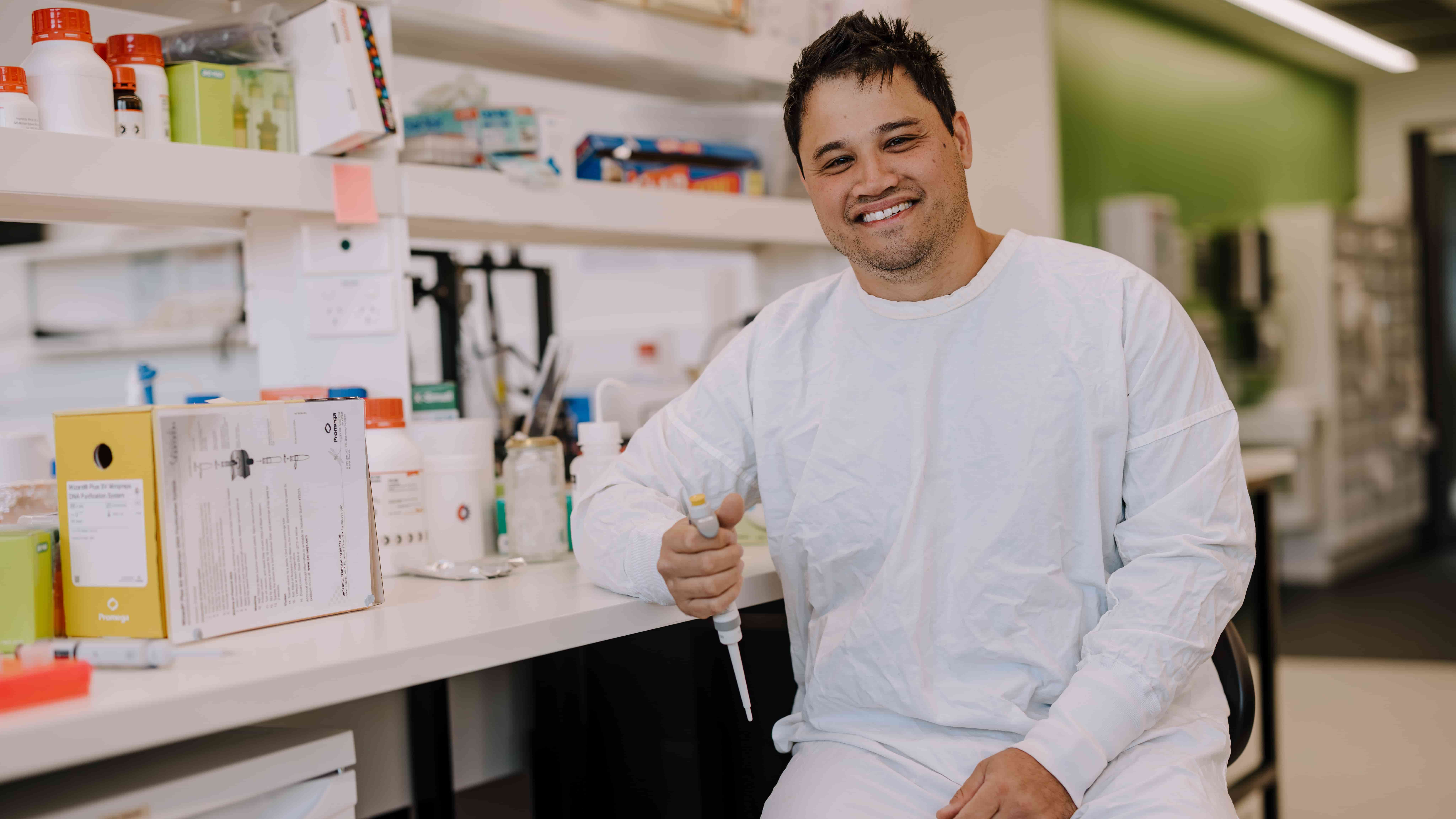 Dr Jeremy Lum wears a white lab coat and sits at a bench in a science laboratory that is covered with equipment. He is smiling. Photo: Michael Gray