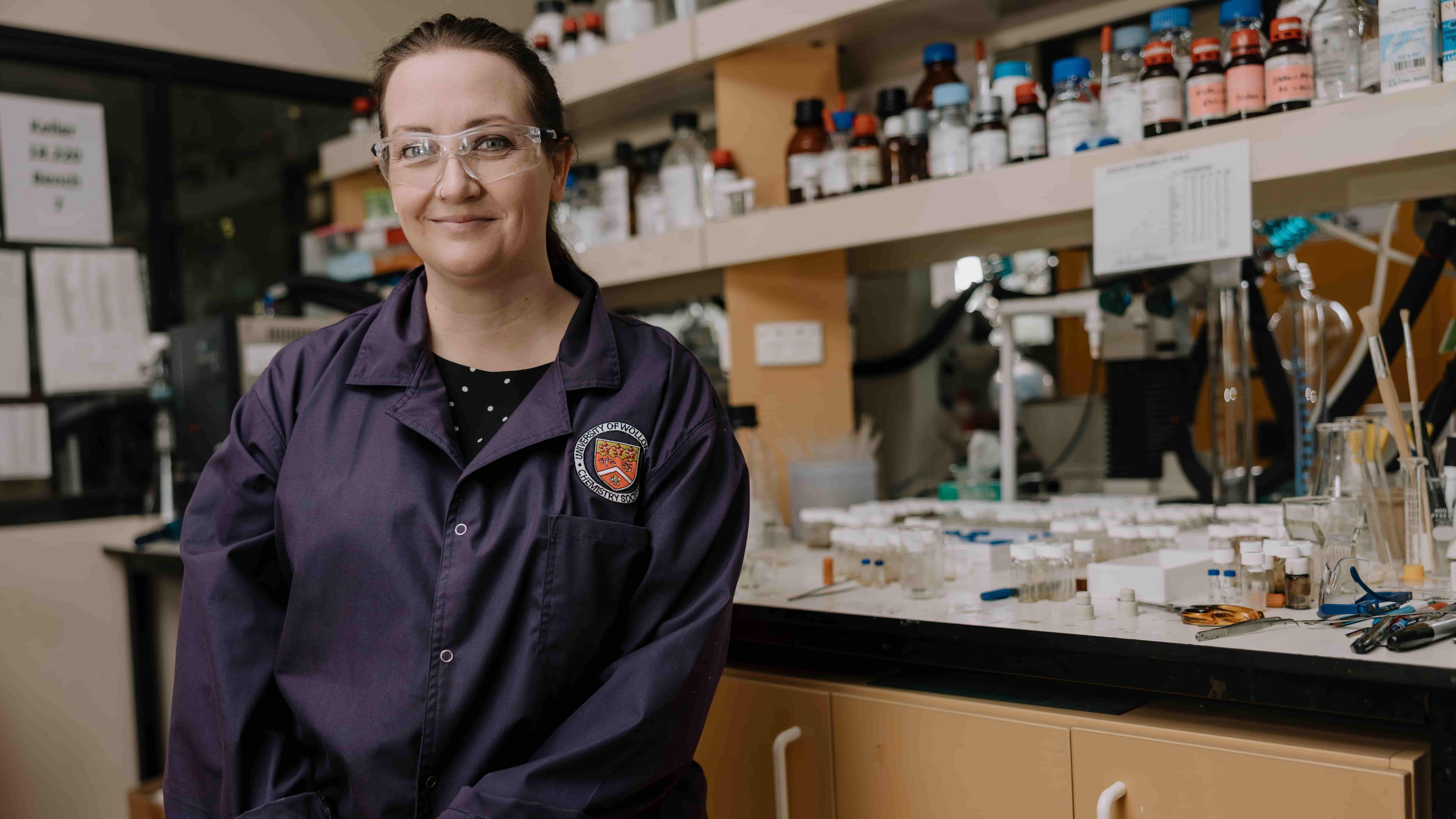 Dr Jennifer Baker sits at a chair at a lab bench, wearing protecting goggles and a dark purple lab coat. On the shelves behind her are vials and chemistry equipment. Photo: Michael Gray