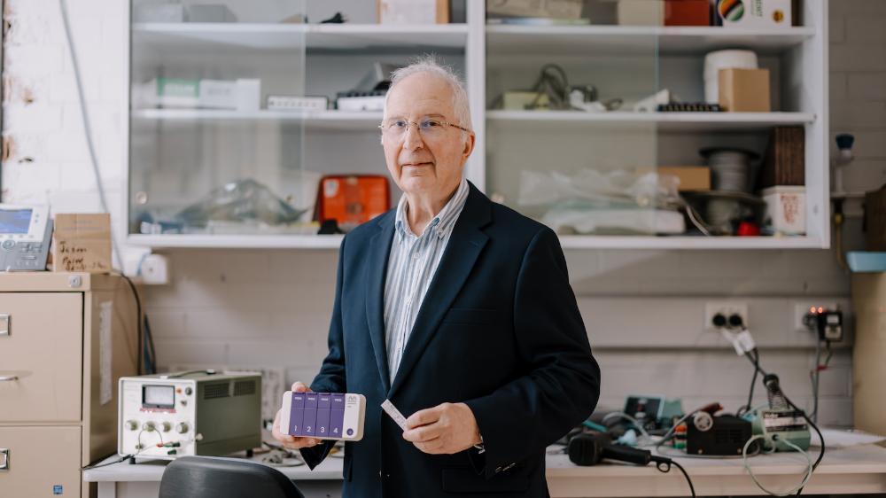 A man stands in a laboratory environment holding a small white and purple device. Photo: Michael Gray