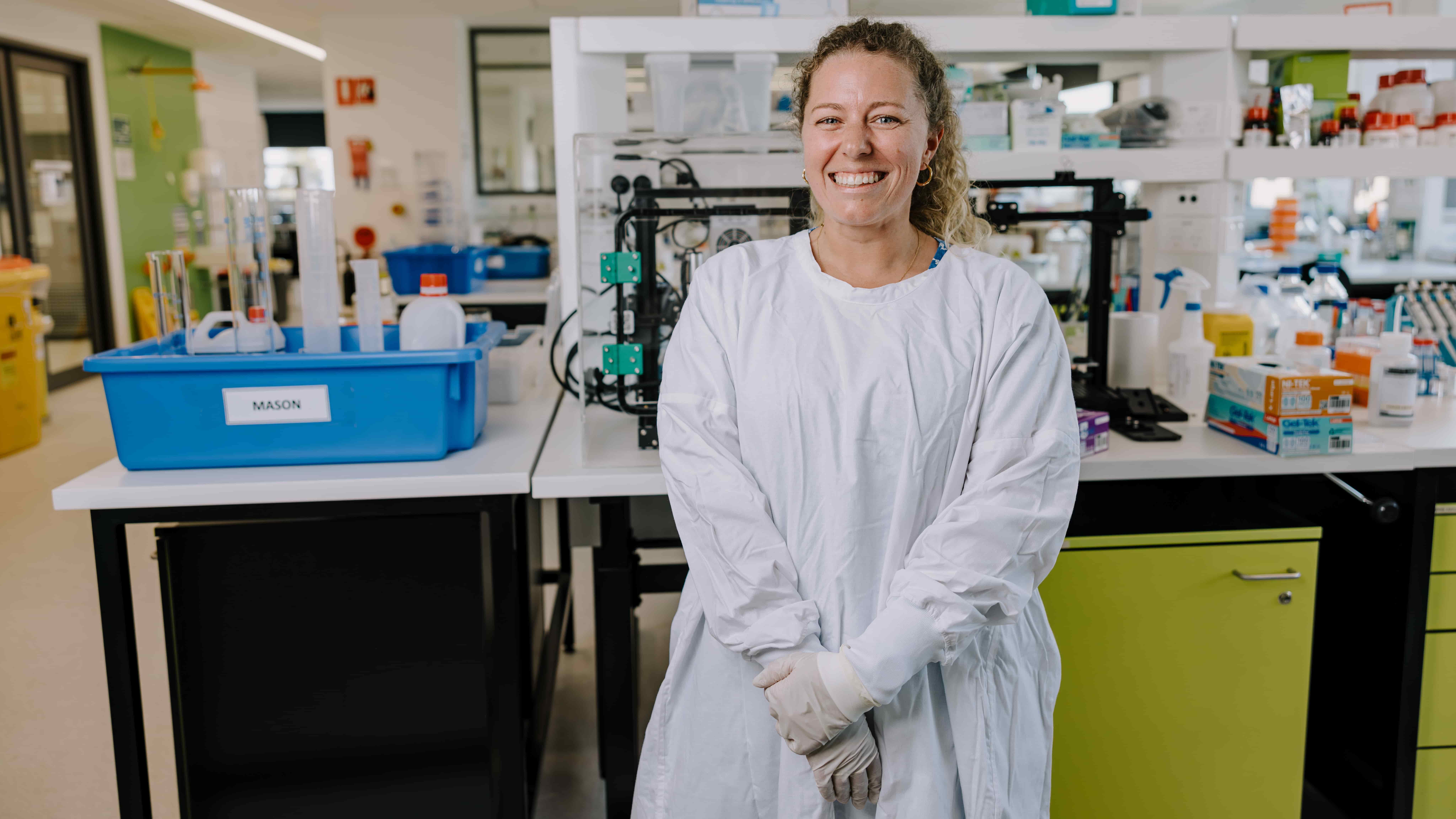 Dr Samara Walpole wears a white lab coat and stands at a science lab with her arms crossed and a smile. Photo: Michael Gray