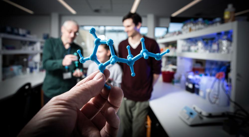 Associate Professor Aaron Oakley holds an example of the 3D chemistry model. Next to him is student Karlee Symonds and Dr Patrick McClosker, who helps Karlee in the lab. They are situated in a laboratory and are all smiling. In the foreground, a hand holds a blue 3D printed chemistry model. Photo: Paul Jones