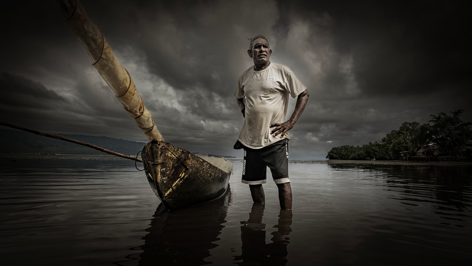 A man stands in the water with his hands on his hips next to a traditional wooden canoe. The sky in the background is dark and stormy. Photo: Paul Jones
