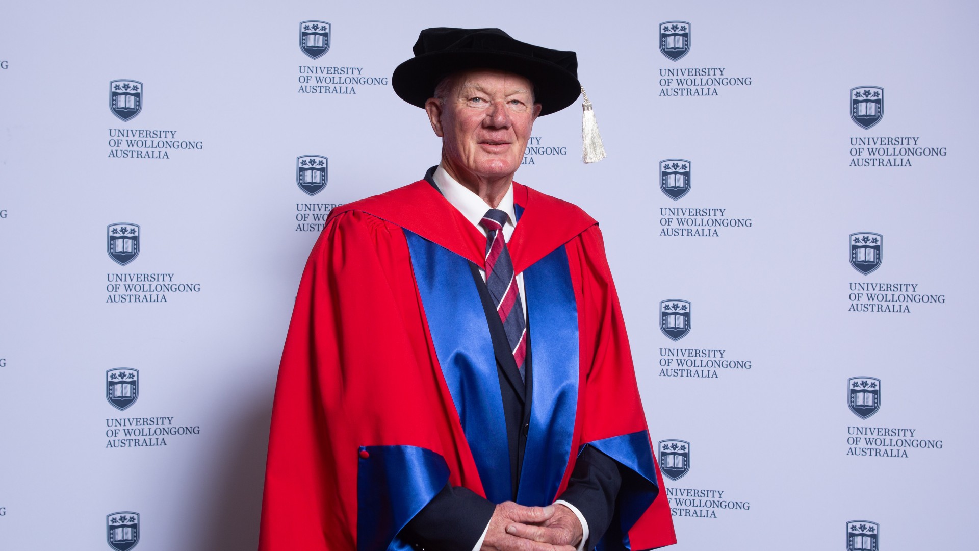 Dr Robert Moses stands in front of a UOW media wall wearing a black and graduation gown and cap. Photo: Andy Zakeli