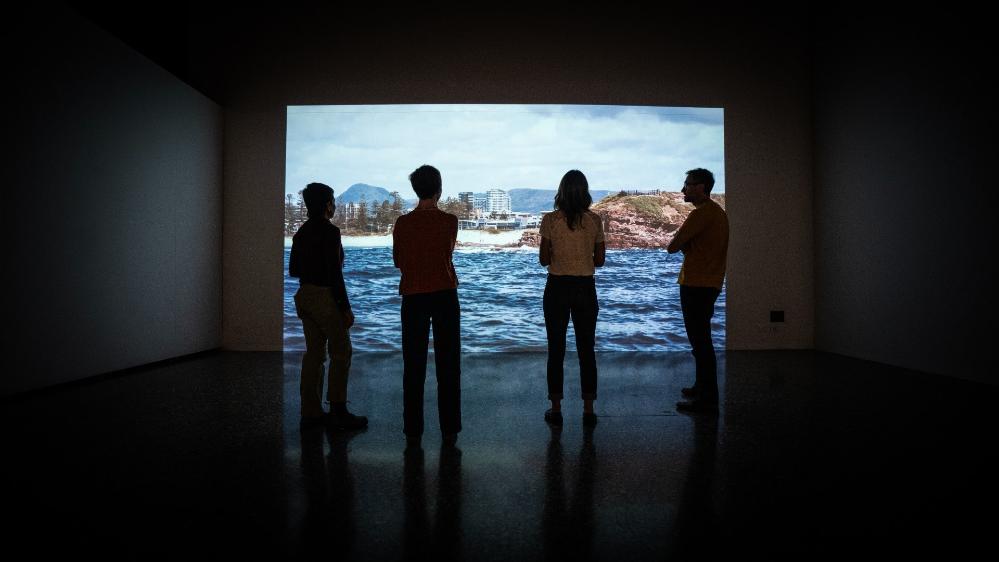 The researchers behind the City+Sea exhibition Kim Williams, Sarah Hamylton, Leah Gibbs, with Lucas Ihlein have their backs to the camera as they watch a video on the screen. . Photo: Paul Jones