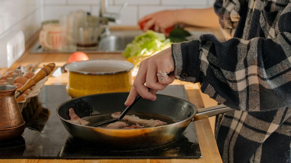 A person cooking bacon in a pan on a stovetop, with nearby eggs, a coffee pot, and fresh vegetables.