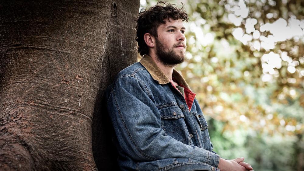 UOW graduate Dakota Feirer sits against a tree and looks into the distance. He wears a denim jacket. Photo: Paul Jones