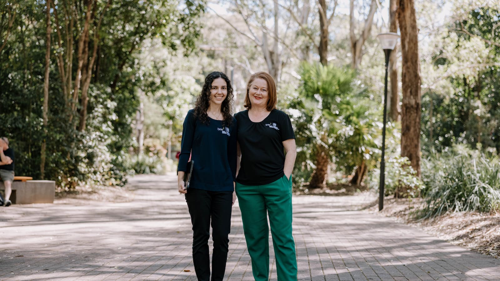 A woman with brown curly hair and a woman with shorter red hair smile as they stand among the trees on Wollongong Campus. Photo: Michael Gray
