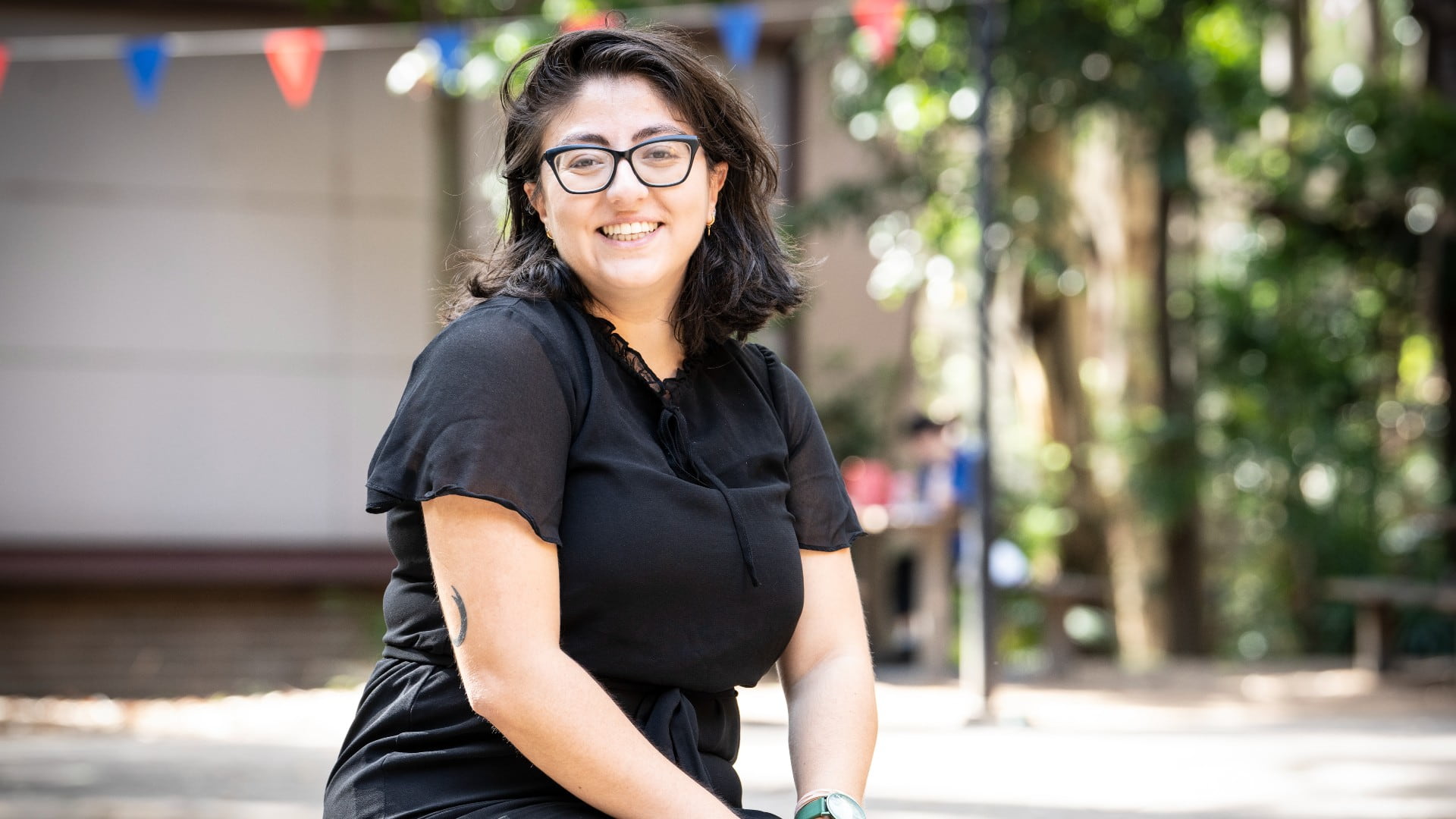 Eda Gunaydin wears a black dress and black glasses, sits on a bench and smiles at the camera. The background is slightly blurry. Photo: Paul Jones