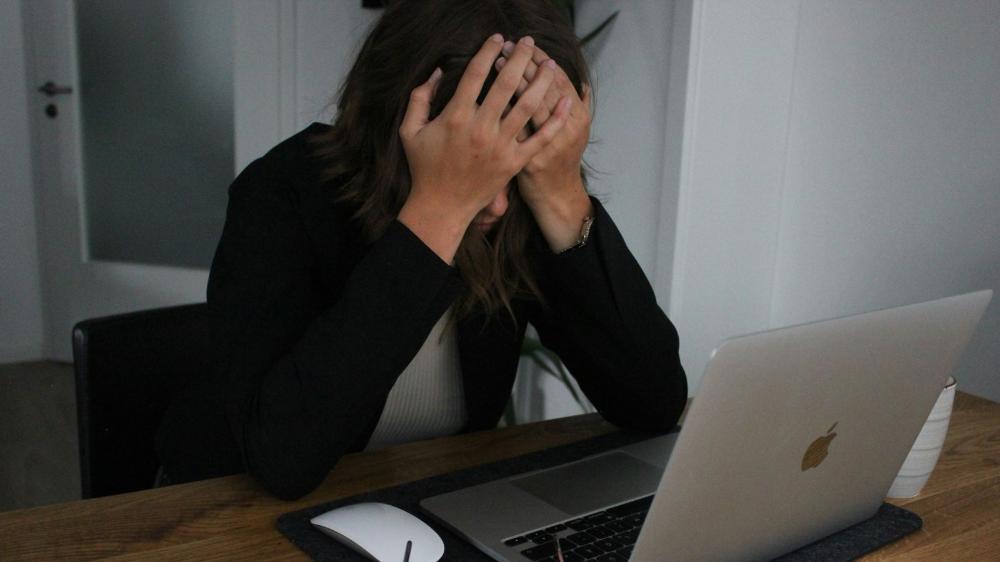 A woman sits at a desk with her head in her hands, looking frustrated