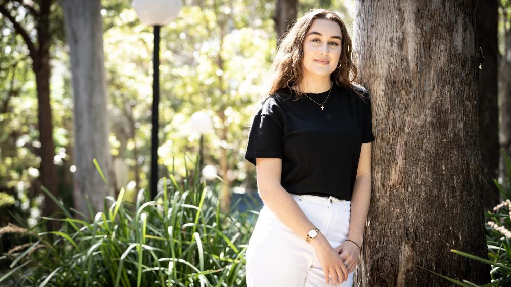 Hannah Lawrence stands against a tree, with a green backdrop. She wears a black shirt and white pants. Photo: Paul Jones