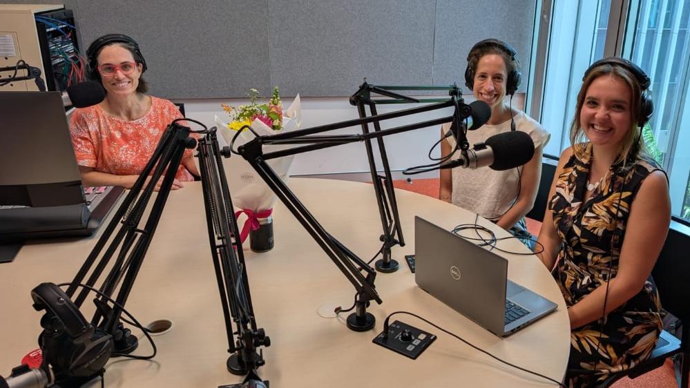 Three women wearing headphones sit around a table in front of microphones.