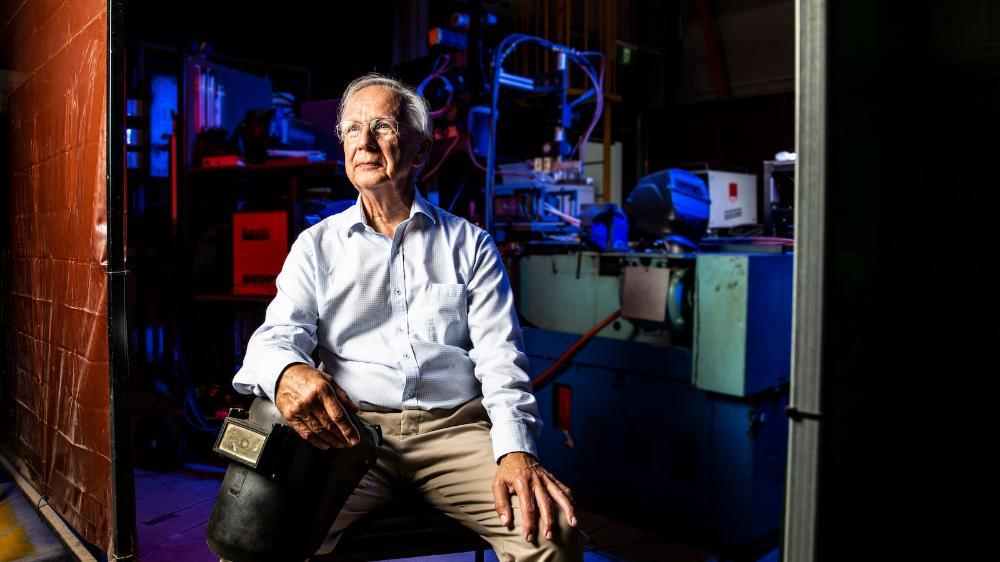 John Norrish sits in a dimly lit industrial workshop, illuminated by blue and red light.