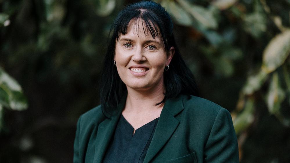 Early childhood educator Karrissa Corsaro stands in front of a green, nature background. She wears a black dress and green jacket and is smiling at the camera. Photo: Michael Gray