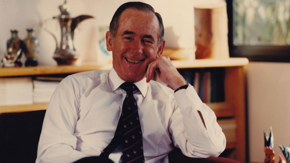 A man sits resting his head on his hand and smiles at the camera