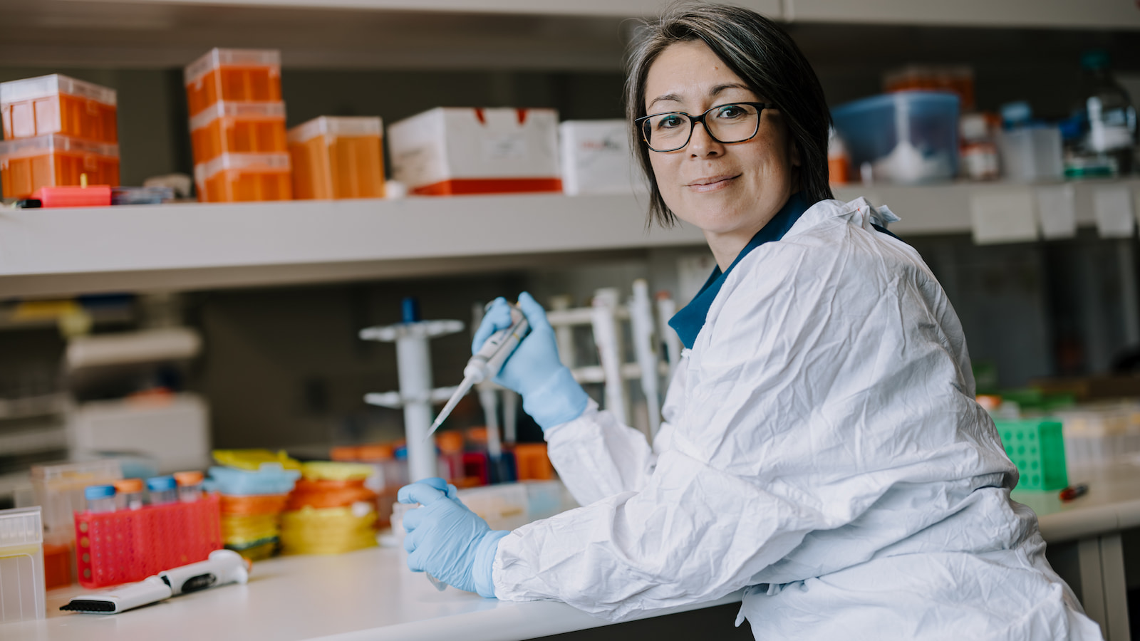Lezanne Ooi sits at a lab bench, wearing a white lab coat. Photo: Michael Gray