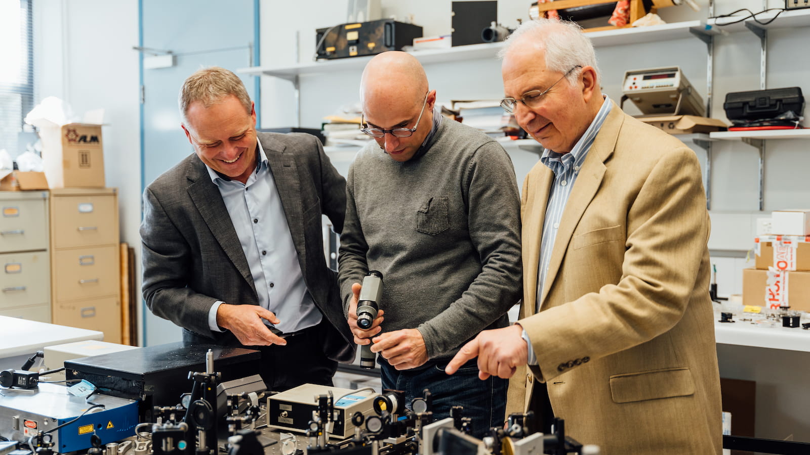 Three men, all smiling and wearing suits, look into and point at a microscope. They are in a science lab.