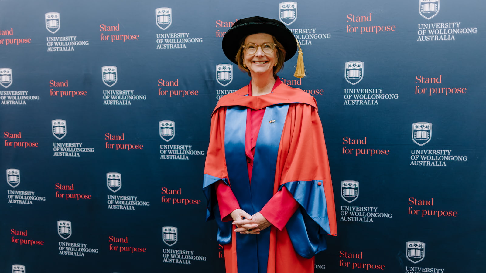 Julia Gillard wears the graduation cap and gown and stands in front of a UOW media wall, smiling. Photo: Michael Gray