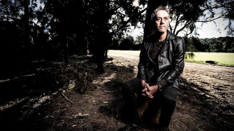 Owen Price wears a leather jacket and sits on a log surrounded by trees in a serene nature setting