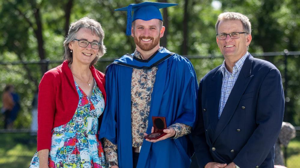 Rhys Smith (centre), wears a blue graduation cap and gown and holds the ý Medal. To his left is his mother. She wears a dress and red cardigan and smiles at the camera. To his right is his father. He wears a blue suit and smiles at the camera. Photo: Andy Zakeli