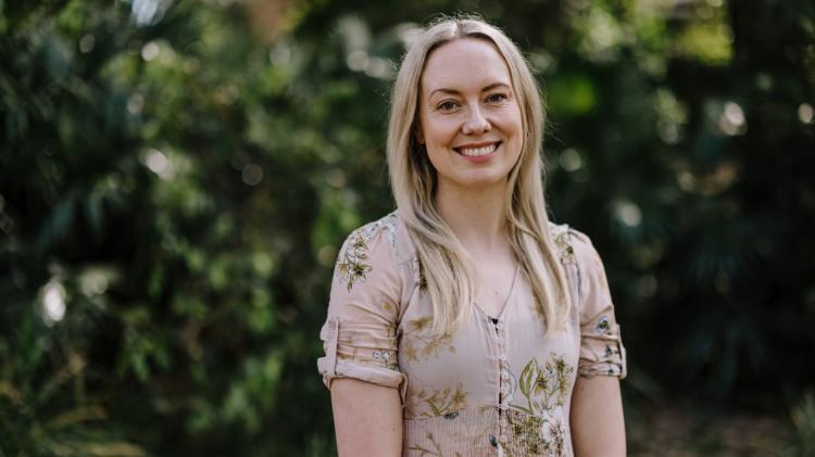 A woman stands looking at the camera smiling, with bushes in the background. She wears a pink coloured dress. Photo: Michael Gray
