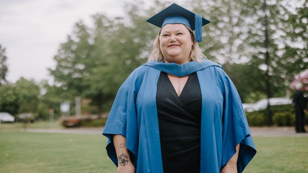 Meg Tattam smiles at the camera and wears a blue graduation gown and cap with a black dress. Photo: Michael Gray