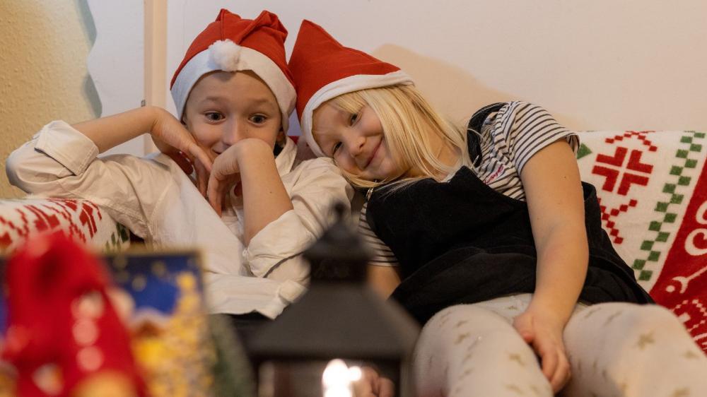 Two children sit together on a sofa and they are wearing Santa hats