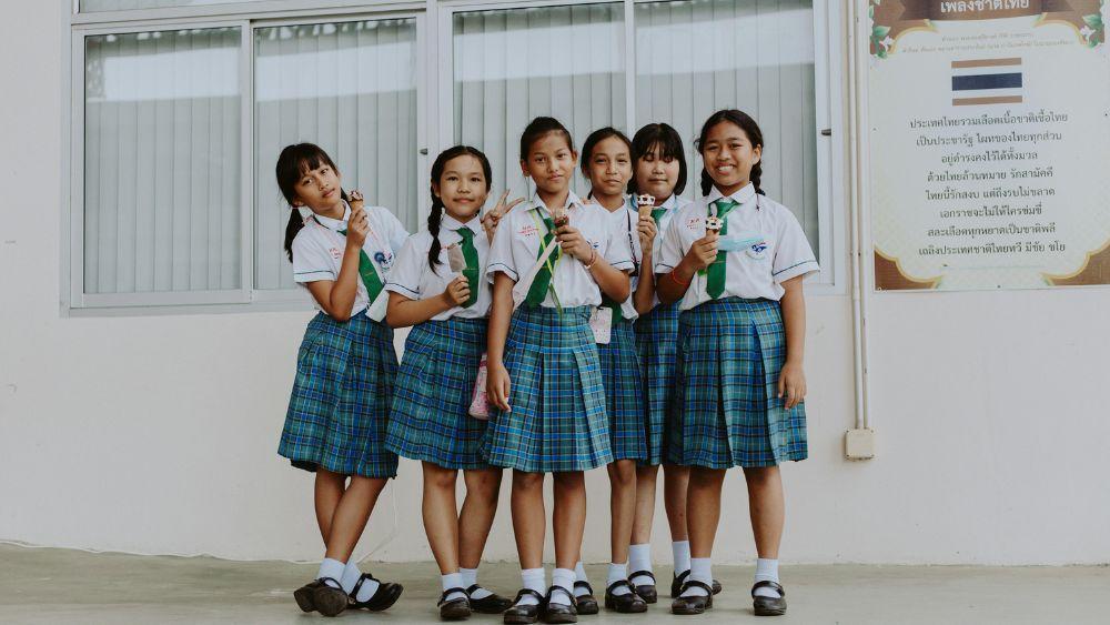 A group of six schoolgirls in uniforms stands together in front of a window, smiling and holding ice cream cones