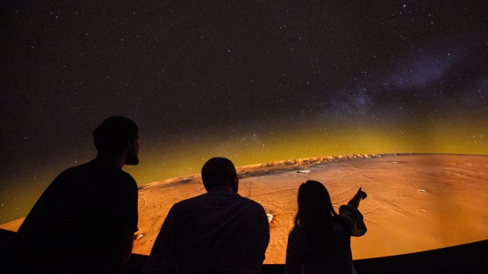 A silhouette of three people looking at an orange hued image of space