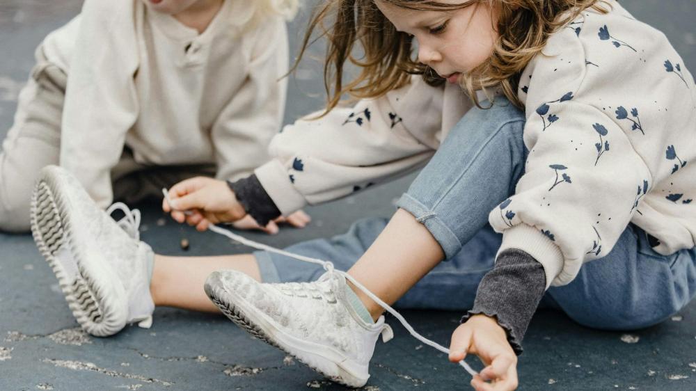 A young child sits on the ground while tying up shoelaces