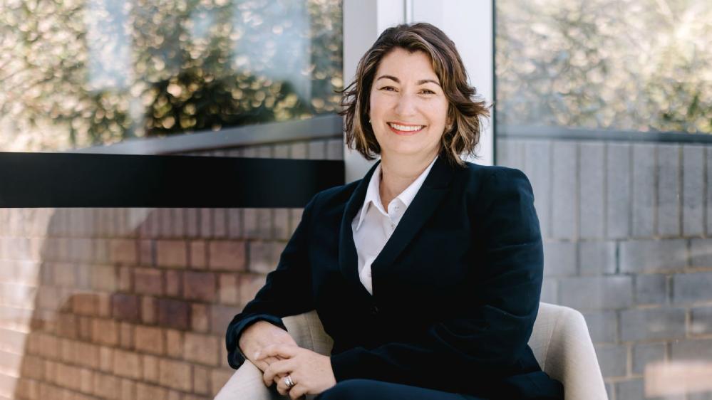 A woman in a black suit is seated in front of a glass window and brick wall and smiling