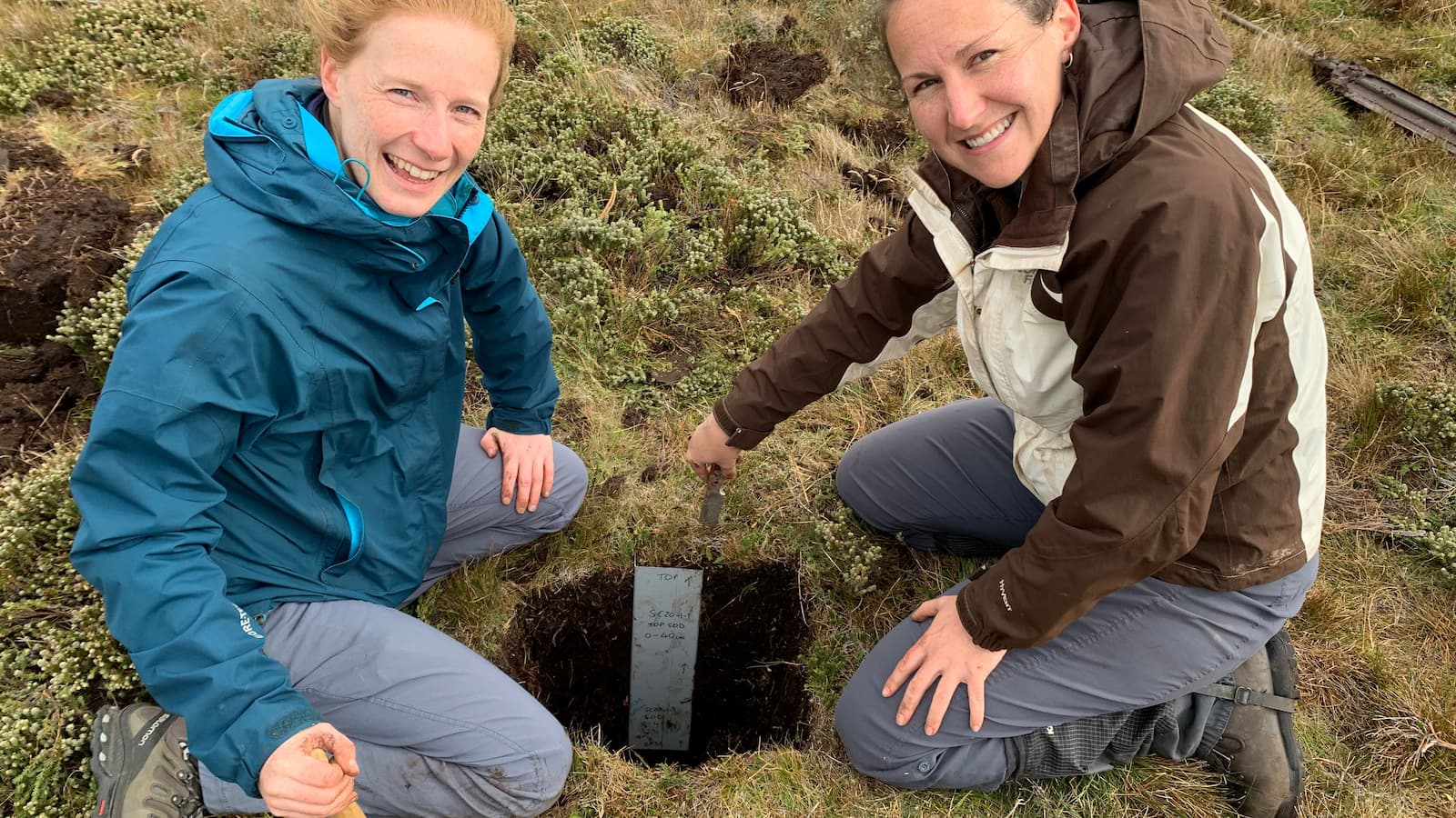 Zoe Thomas and Haidee Cadd kneel in the grass while extracting peat bogs in the Falkland Islands. Photo: Christian Turney