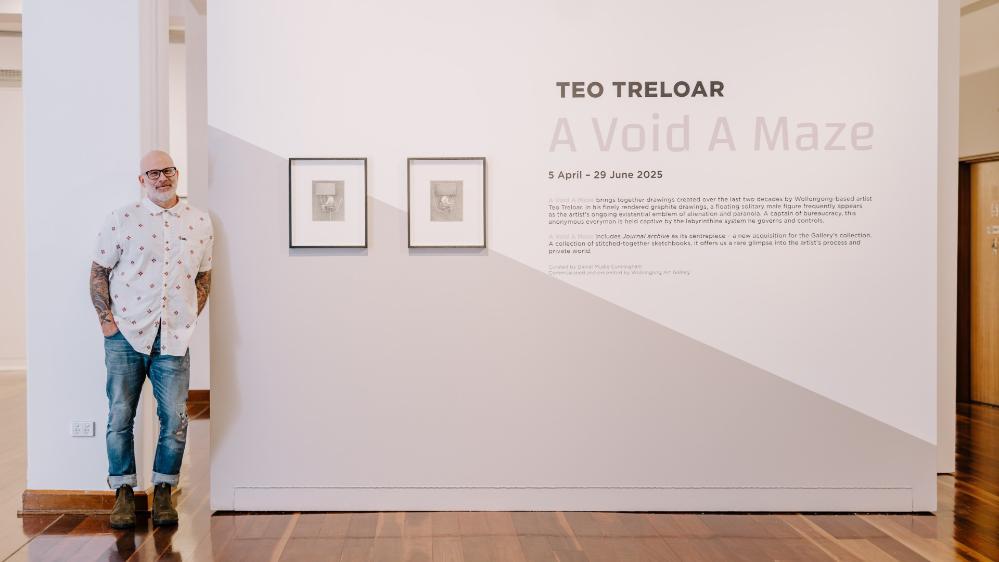 A man stands in a light-filled gallery with framed pictures on the wall behind him. Photo: Michael Gray