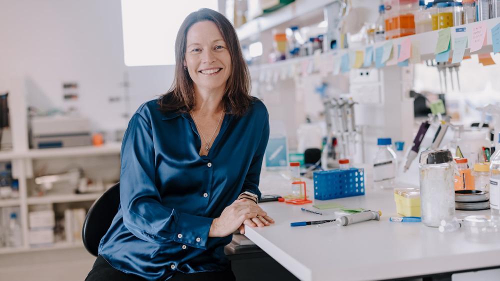 A female wearing a blue shirt sits at a laboratory desk, surrounded by test tubes and science items. Photo: Michael Gray