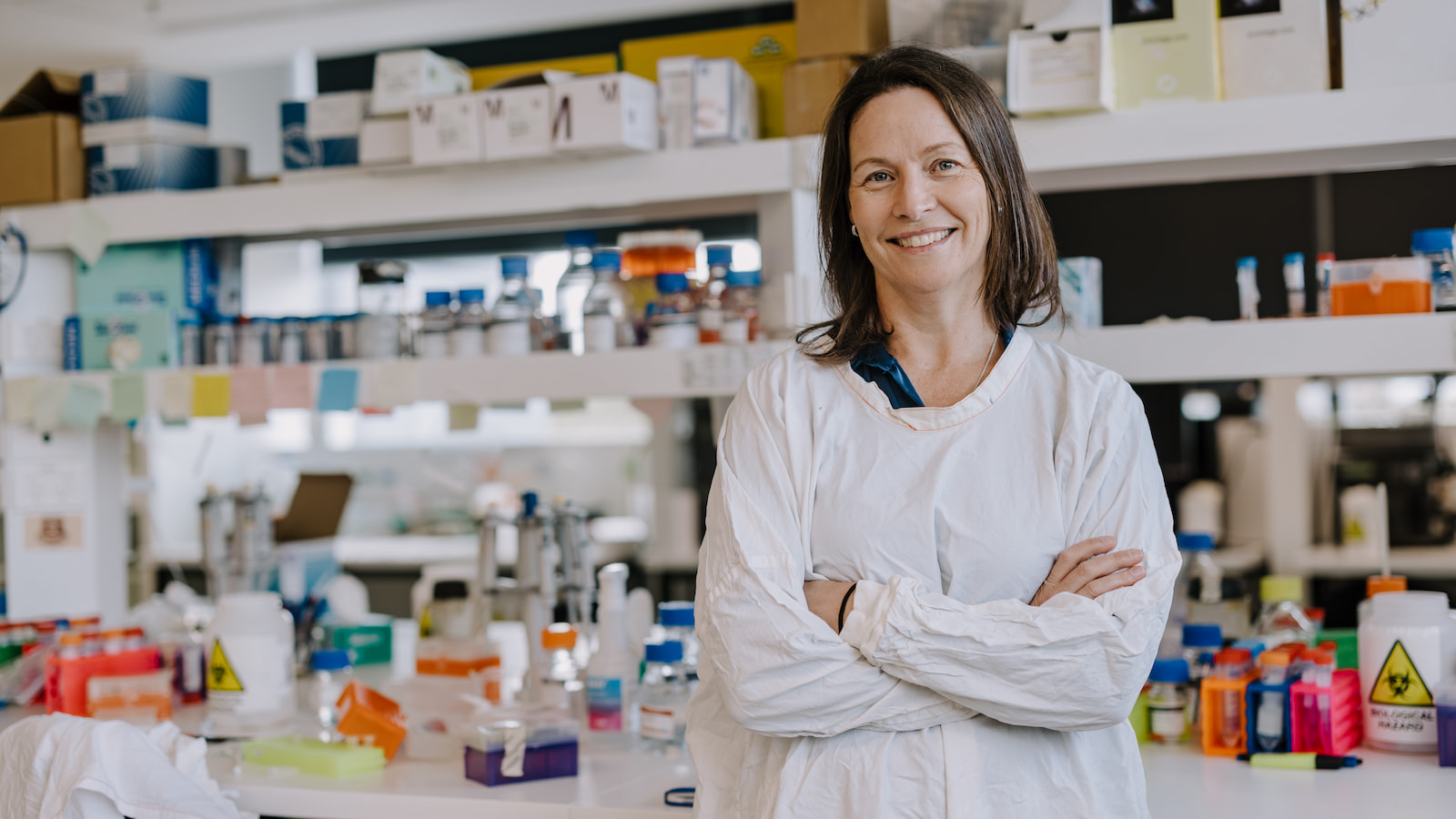 Christen Chisholm wears a white lab coat with her arms crossed as she stands at a lab bench. Photo: Michael Gray