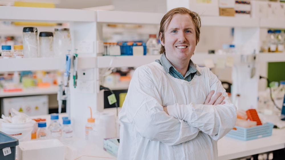 A male wearing a white lab coat is smiling in the research lab, with his arms crossed. Photo: Michael Gray