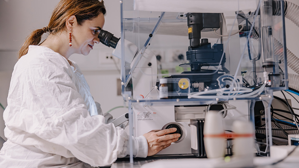 A researcher dressed in a lab coat looks through a microscope.