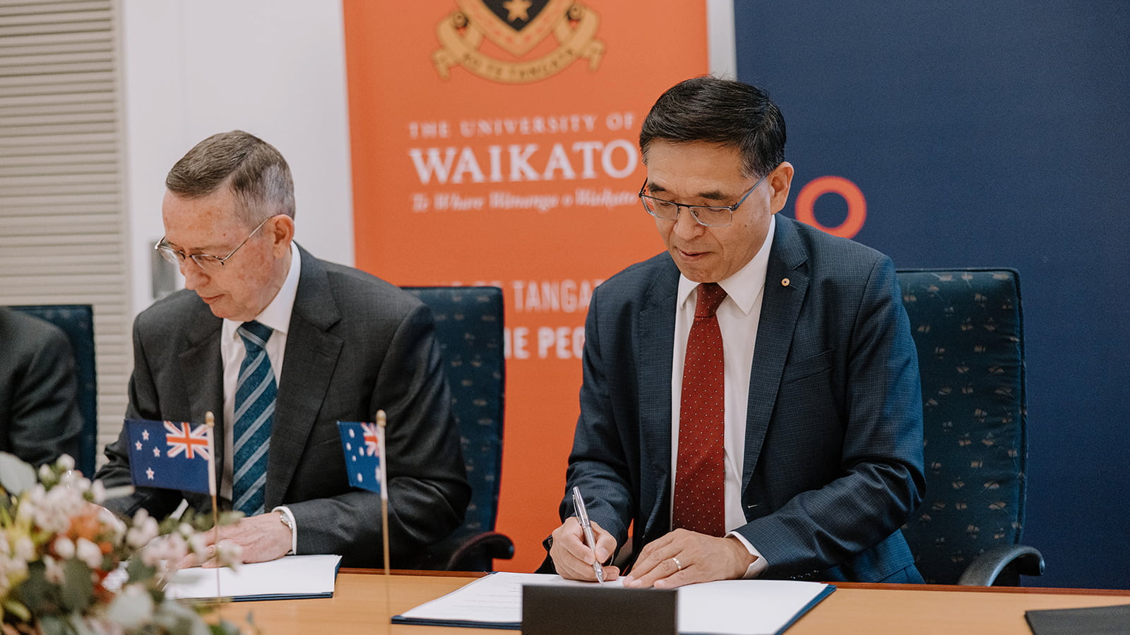 Two men sit at a table in a boardroom and sign a folder in front of them. Photo: Michael Gray