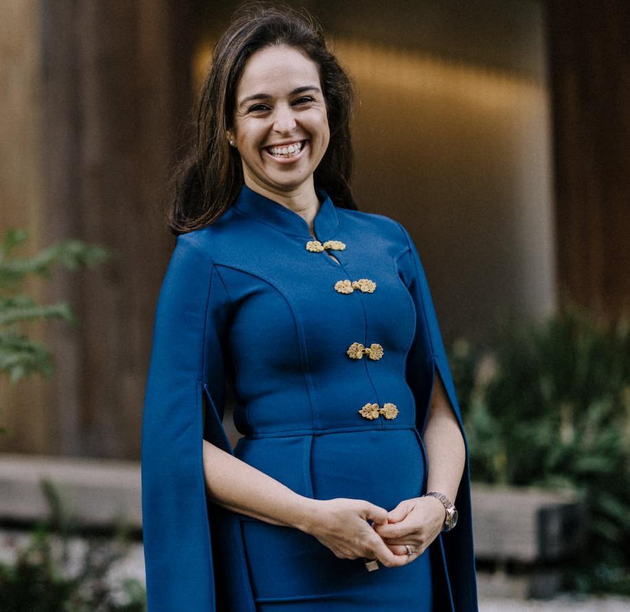 Dr Izabela Pereira Watts, wearing a blue dress with gold buttons, stands in front of a building smiling at the camera.