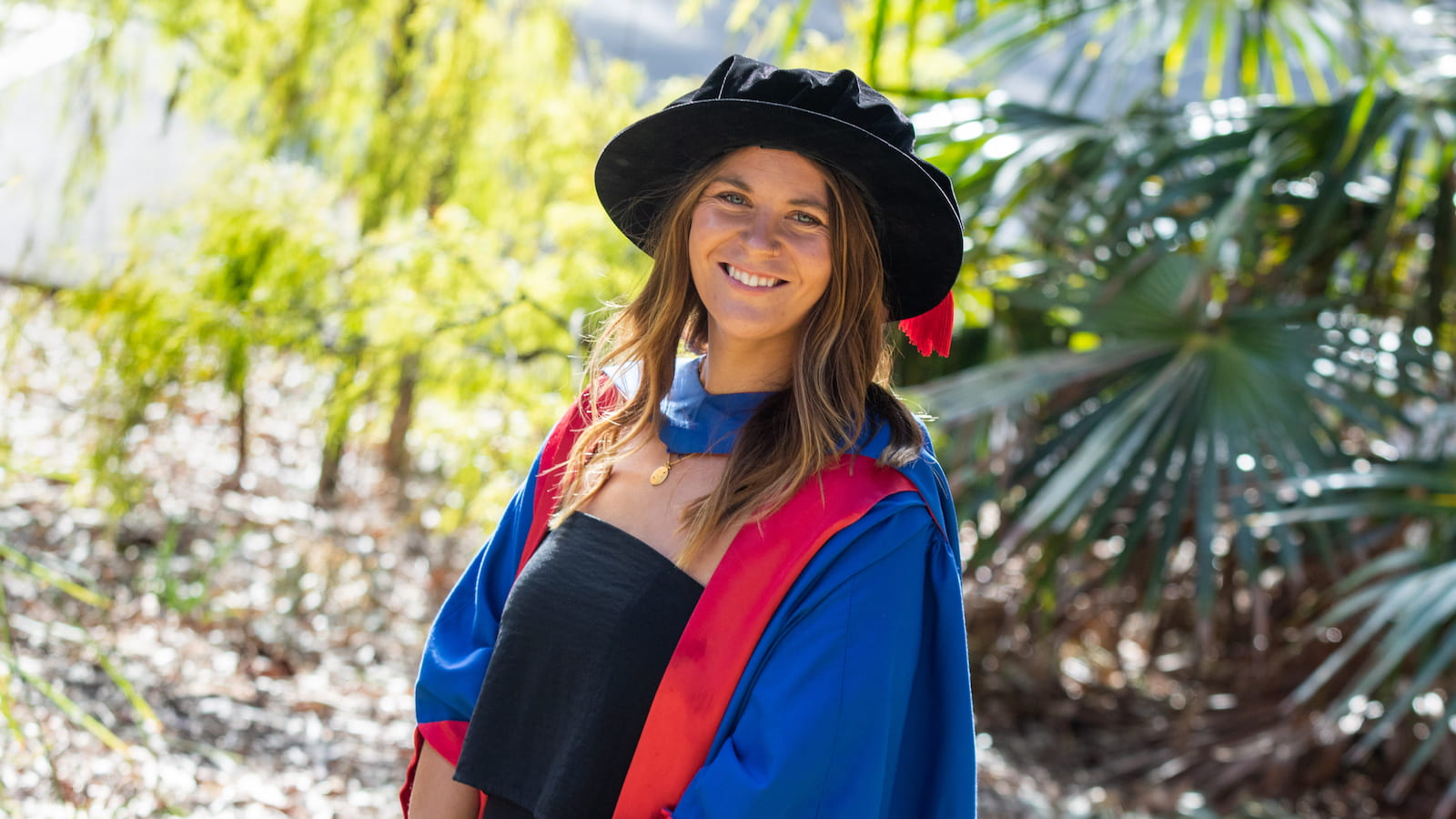 Dr Freya Croft, wearing a black dress, blue and red graduation gown, and black cap, smiles at the camera. There are green bushes in the background. Photo: Andy Zakeli