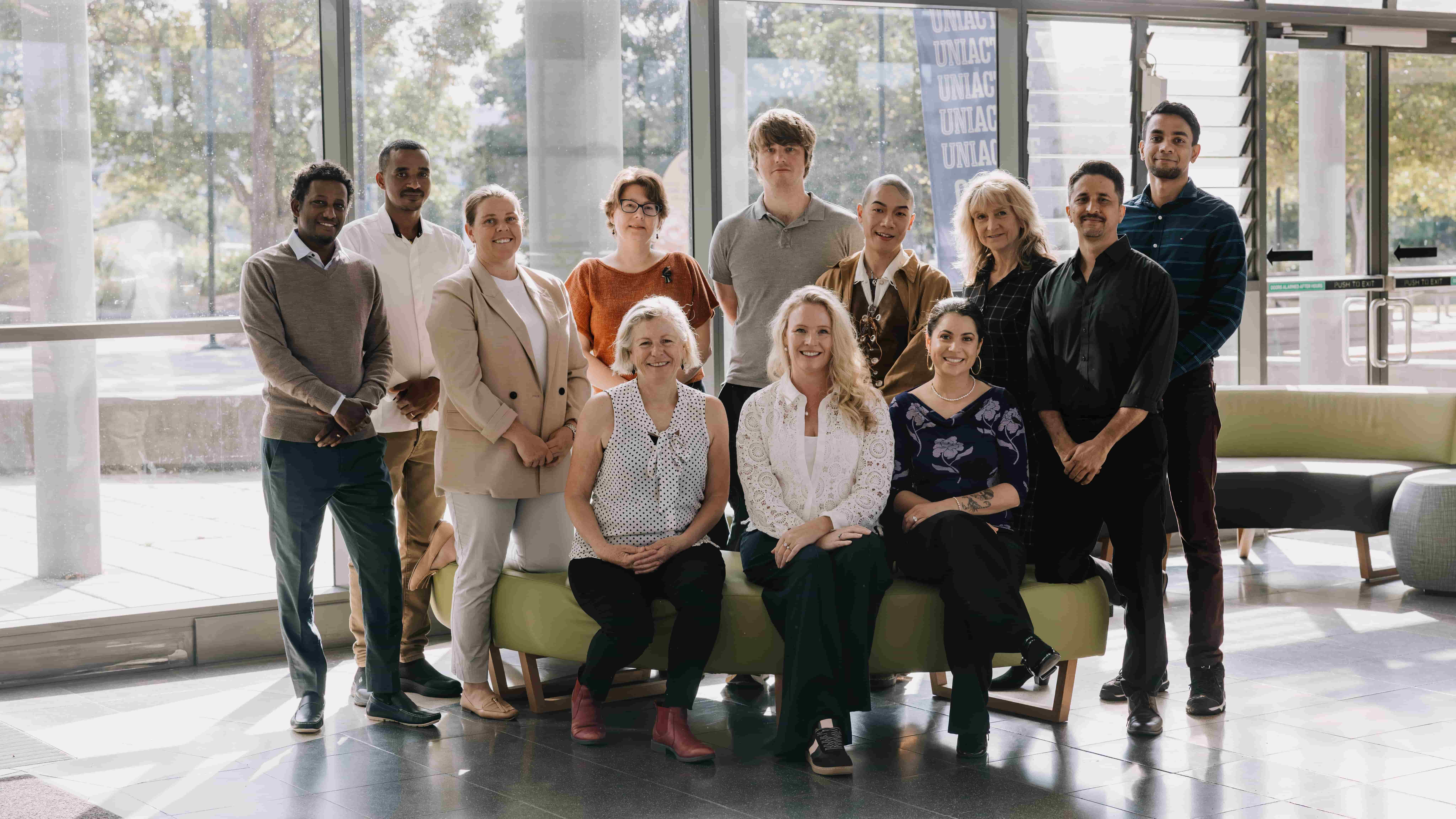 A team of researchers from the Palliative Care Outcomes Collaboration, including Director Sabina Clapham (front centre), are arranged around a lounge in the foyer of a building. Three people are seated and nine people stand behind. Photo: Michael Gray